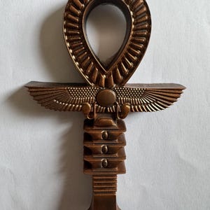 May include: Bronze-colored Ankh pendant. The pendant features a teardrop-shaped loop at the top, a horizontal bar with stylized wings, and a vertical base with stacked rectangular elements. The Ankh is a symbol of life and immortality.