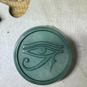 May include: A round, teal-colored pendant featuring the Eye of Horus symbol. The pendant has a raised border and a detailed depiction of the ancient Egyptian symbol. The pendant is set against a neutral, textured background.