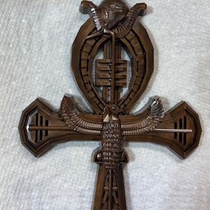 May include: A dark brown, carved wall ornament shaped like an Egyptian ankh cross. The design features a central falcon with outstretched wings, an Eye of Horus at the top, and intricate geometric patterns throughout the structure.