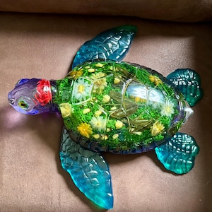 May include: A decorative turtle figurine with a clear, textured shell filled with green and yellow floral elements. The turtle's head is purple and red, and its flippers are a translucent blue-green color. The figurine is resting on a brown surface.