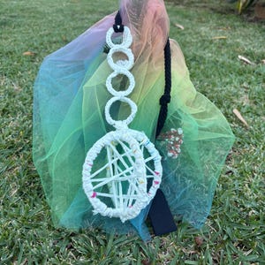 Peut inclure: Suspension murale en macramé blanc avec un motif circulaire et un cordon noir. Le macramé est présenté sur un fond de tissu de tulle aux couleurs de l'arc-en-ciel. L'ensemble est posé sur une pelouse verte.