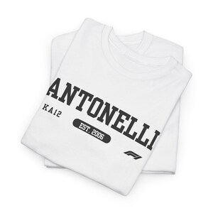 May include: A white short-sleeved t-shirt featuring black text that reads ANTONELLI, KA12, and EST. 2006, along with a small black Formula 1 logo. The shirt is folded neatly, displaying the graphic print clearly on the front chest area.