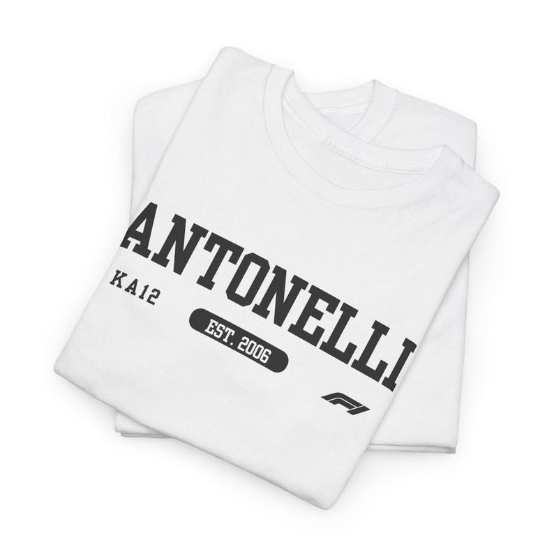 May include: A white short-sleeved t-shirt featuring black text that reads ANTONELLI, KA12, and EST. 2006, along with a small black Formula 1 logo. The shirt is folded neatly, displaying the graphic print clearly on the front chest area.