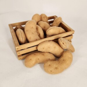 Könnte beinhalten: Eine Holzkiste gefüllt mit mehreren hellbraunen, plüschigen, kartoffelförmigen Spielzeugen. Einige Kartoffeln quellen aus der Kiste. Die Kartoffeln haben kleine, dunkelbraune, aufgenähte Details, die Kartoffelaugen ähneln.