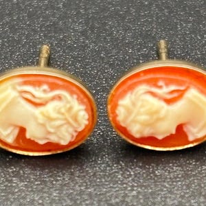 May include: A pair of oval-shaped cameo earrings. Each earring features a cream-colored profile set against an orange background, framed by a gold-colored metal. The earrings have a post for pierced ears.