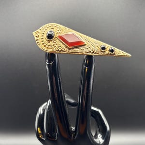 May include: A gold-toned brooch with an elongated, geometric design. It features a central red gemstone and smaller black stones, with intricate embossed detailing. The brooch is held up by a black hand.