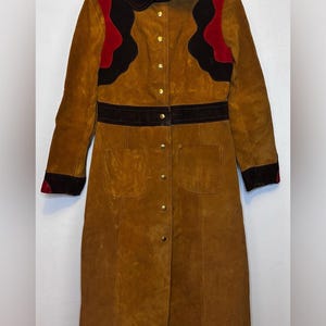 May include: A long tan suede coat featuring a dark brown collar, cuffs, and waist band. The upper chest area displays scalloped dark brown and red suede panels. The coat has a front button closure with gold-toned metal buttons and two pockets.