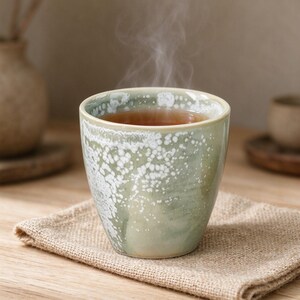 May include: A ceramic cup with a tapered shape and a reactive glaze finish in shades of sage green and white. The cup is filled with a warm brown liquid, and visible steam rises from the top. The exterior features a mottled, speckled texture.