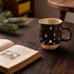 May include: A ceramic mug with a black exterior featuring gold-colored star patterns and white mountain silhouettes at the base. The mug has a glossy finish, a curved handle, and a light beige interior. It is positioned next to an open book.