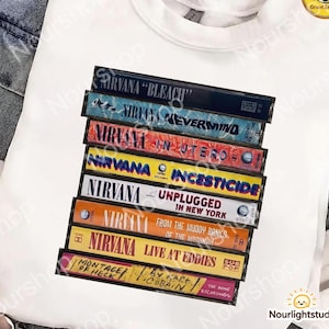 May include: A white t-shirt featuring a graphic print of a stack of eight Nirvana cassette tapes. The tapes are labeled Bleach, Nevermind, In Utero, Incesticide, Unplugged in New York, From the Muddy Banks of the Wishkah, Live at Eddies, and Montage of Heck.
