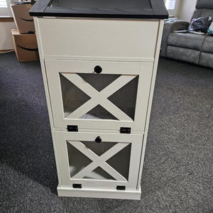 May include: A tall, off-white storage cabinet with a black top and mesh panel. The cabinet has two lower compartments with an X-shaped design and mesh inserts. Black hardware and a black knob complete the look. The cabinet is on a gray carpet.