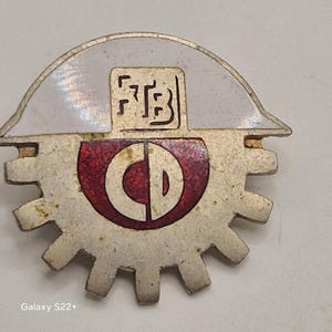 Könnte beinhalten: Eine Vintage-Metallnadel mit einem zahnradförmigen Sockel und einem weißen und roten Emblem. Das Emblem zeigt die Buchstaben "FTB" und "CD". Die Nadel hat eine silberne Oberfläche und eine weiße, gebogene Oberseite.