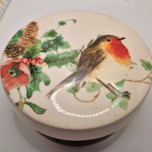 May include: A round, off-white ceramic trinket box with a lid featuring a detailed illustration of a robin bird with a red breast, perched on a branch with green leaves and red berries. The lid also has a pine cone and a red flower.