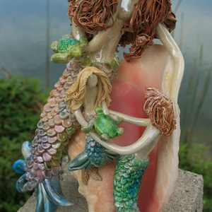 May include: A ceramic sculpture of three mermaids sitting on a large pink seashell. The mermaids have long flowing hair and iridescent tails. The mermaids are holding small green sea turtles.