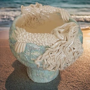 May include: A ceramic bowl featuring a textured, light blue and white mottled finish. The exterior is adorned with sculpted, off-white relief figures of mermaids with detailed scales and flowing hair that wrap around the rim of the vessel.
