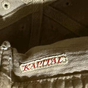 May include: Close-up of the inside of a tan baseball cap. The cap features a red embroidered "KAPITAL" patch. The cap's interior has a textured, woven design and visible stitching.