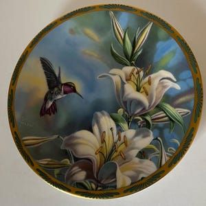 May include: Decorative plate featuring a hummingbird in flight near white lilies. The plate has a gold rim with a green and gold border. The background is a soft blue and green.