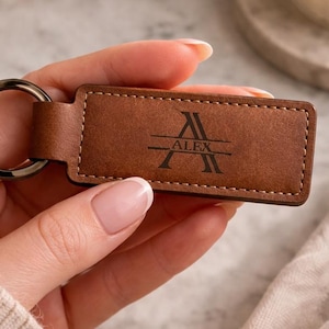 May include: A brown leather keychain with a silver ring and clasp. The rectangular tag is embossed with the name "ALEX" and a stylized letter "A". The keychain is held in a hand, with a blurred background including a candle and decorative elements.