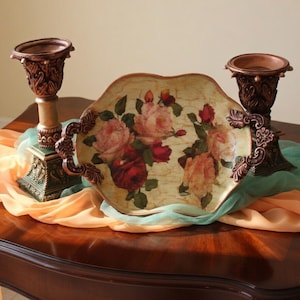May include: A decorative oval tray with scalloped edges featuring a floral print of pink, red, and yellow roses on a cream background, flanked by two ornate, dark bronze-colored candle holders with intricate relief detailing and flared rims.
