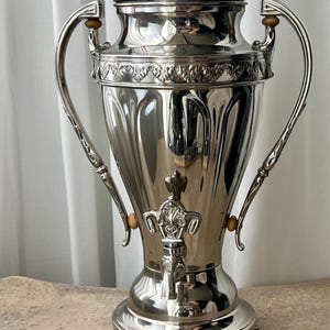 May include: A polished silver coffee urn with a clear glass finial on the lid. The urn has two curved handles and a decorative spigot. The design includes ornate detailing around the top and base, reflecting light.