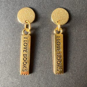May include: A pair of gold-coloured earrings. Each earring has a small, textured, circular stud connected to a rectangular charm. The charm is inscribed with the words "I LOVE BOOKS".