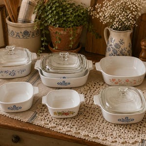 May include: A collection of vintage white and glass CorningWare dishes with floral and blue patterns. The set includes various sizes of square and rectangular baking dishes, some with lids. The dishes are arranged on a wooden surface.