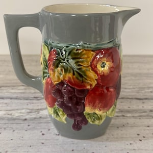 May include: Gray ceramic pitcher with a handle and spout. The pitcher is decorated with a relief pattern of colorful fruits, including red apples, purple grapes, and green leaves. The interior is a cream color.
