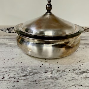 May include: A silver-colored, lidded serving dish with a dark brown finial. The dish has a rounded base and a lid with a decorative handle. The dish has ornate handles on either side. The dish is likely made of metal.