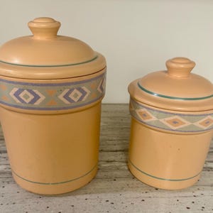May include: Two peach-colored ceramic canisters with lids. The canisters have a decorative band of geometric shapes in blue, lavender, and orange. The lids have a small knob on top and a thin green line around the edge. The canisters are different sizes.