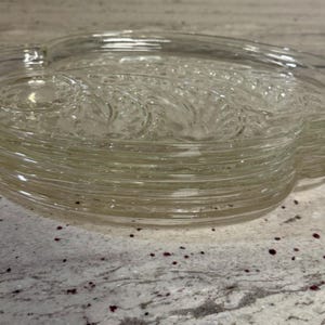 Pode incluir: Uma pilha de travessas de vidro transparente. Cada travessa tem um design em forma de coração com uma borda ondulada e um padrão em relevo detalhado. As travessas estão empilhadas em uma bancada salpicada.