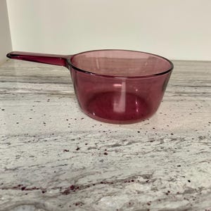 May include: A clear, burgundy-colored saucepan with a long handle. The pan is made of glass and sits on a light-colored countertop. The handle extends to the left, and the pan's bowl is round.