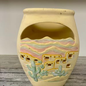 Puede incluir: Jarrón de cerámica color crema con una gran abertura arqueada. El jarrón presenta una escena del desierto del suroeste con montañas en colores pastel, cactus y edificios de estilo pueblo. El diseño está tallado y pintado.