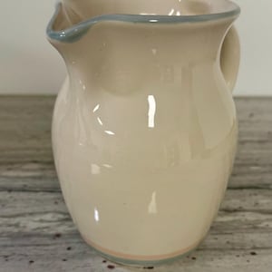 May include: Cream ceramic jug with a light blue rim and handle. A thin pink and blue band encircles the base. The jug has a classic shape and a glossy finish, suitable for serving beverages.