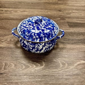 May include: A blue and white enamelware pot with a lid. The pot has two handles and a silver rim. The blue and white pattern is a speckled design. The pot is sitting on a wooden surface.