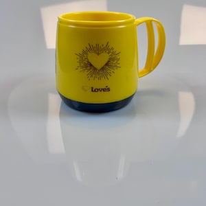May include: Yellow mug with a dark blue base and handle. The mug features a heart design with radiating lines and the word "Love's" printed below the heart. The mug is made of a glossy material.