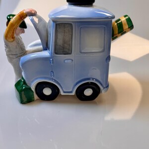 May include: A light blue ceramic teapot shaped like a vintage truck with a black top and black wheels. A figure in a green hat and white overalls appears to be working on the truck. The spout is a green and yellow striped awning.