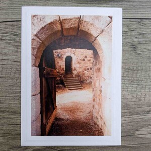 May include: A view through a stone archway, revealing a second archway and a set of stairs leading up to a closed door. The stone walls are a warm brown color, and the ground is covered in dirt and leaves.