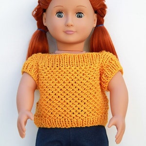 May include: A doll with long ginger hair styled in two pigtails, wearing a short-sleeved, mustard yellow knitted top with a textured diamond pattern and a ribbed neckline, paired with dark blue denim pants against a plain white background.