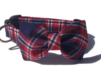 May include: A pet collar featuring a red, navy blue, and white plaid pattern with an attached fabric bow tie. The collar includes a black plastic side-release buckle and a silver-toned metal D-ring for attaching a leash or identification tags.