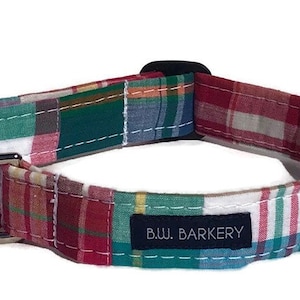 May include: A colorful plaid dog collar with red, green, blue, and white squares. The collar has a black buckle and a silver metal D-ring. A small navy blue tag reads "B.W. Barkery".