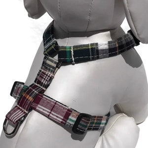 May include: A plaid dog harness in shades of green, burgundy, and white. The harness features adjustable straps and black plastic buckles for a secure fit. A metal D-ring is attached for leash attachment. The harness is designed to fit around the dog's chest and neck.