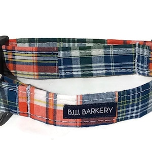 May include: A blue, orange, and white plaid dog collar with a black buckle and silver D-ring. The collar has the text "B.W. BARKERY" stitched on it.