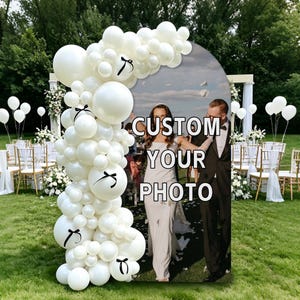 Könnte beinhalten: Ein bogenförmiger Hintergrund mit einem benutzerdefinierten Fotodruck und dem Text CUSTOM YOUR PHOTO, verziert mit einer großen Ansammlung weißer Luftballons in verschiedenen Größen, teilweise mit kleinen schwarzen Schleifen, auf einer Wiese.