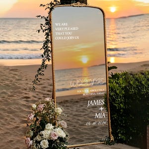May include: A gold-framed mirror with the text "WELCOME" and "YOU LOOK ABSOLUTELY BEAUTIFUL TODAY" is displayed on a sandy beach. The mirror reflects a sunset over the ocean. The names "JAMES + MIA" and the date "04.16.2026" are also visible.