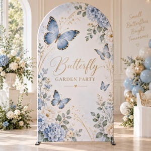 Peut inclure: Un support de toile de fond arqué avec une housse en tissu blanc imprimée de papillons bleus, du texte doré Butterfly GARDEN PARTY, et des illustrations d'hortensias bleus et blancs avec du feuillage vert et des détails dorés.