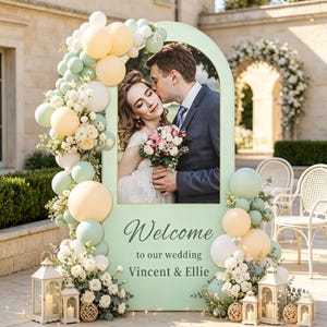 Peut inclure: Un panneau d'accueil de mariage arqué vert clair avec une photo d'un couple, orné de grappes de ballons pastel pêche, blancs et vert sauge et de compositions florales blanches, avec le texte Welcome to our wedding Vincent & Ellie.
