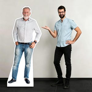 Puede incluir: Un recorte de cartón a tamaño real de un hombre con una camisa blanca, vaqueros azules y zapatos negros. Un segundo hombre con una camisa azul claro, pantalones negros y gafas está de pie junto al recorte, gesticulando con la mano. El fondo es una pared blanca lisa.
