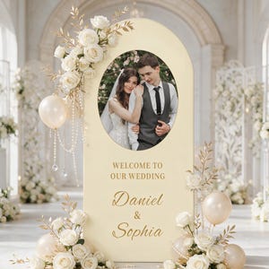 May include: A cream-colored arched wedding welcome sign featuring a central oval photo of a couple, gold text reading "WELCOME TO OUR WEDDING" and "Daniel & Sophia," decorated with white roses, gold leaves, pearl strands, and pale balloons.