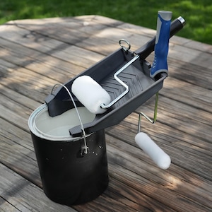 May include: A black paint can with white paint inside, a dark gray paint tray, and two white paint rollers. The paint tray has a blue and gray handle. The items are on a weathered wooden surface.
