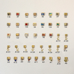 May include: A selection of gold-coloured charms featuring diverse designs such as letters, hearts, and symbols. Each charm is rectangular or square, with a small loop for attachment. Numbers are printed on each charm.